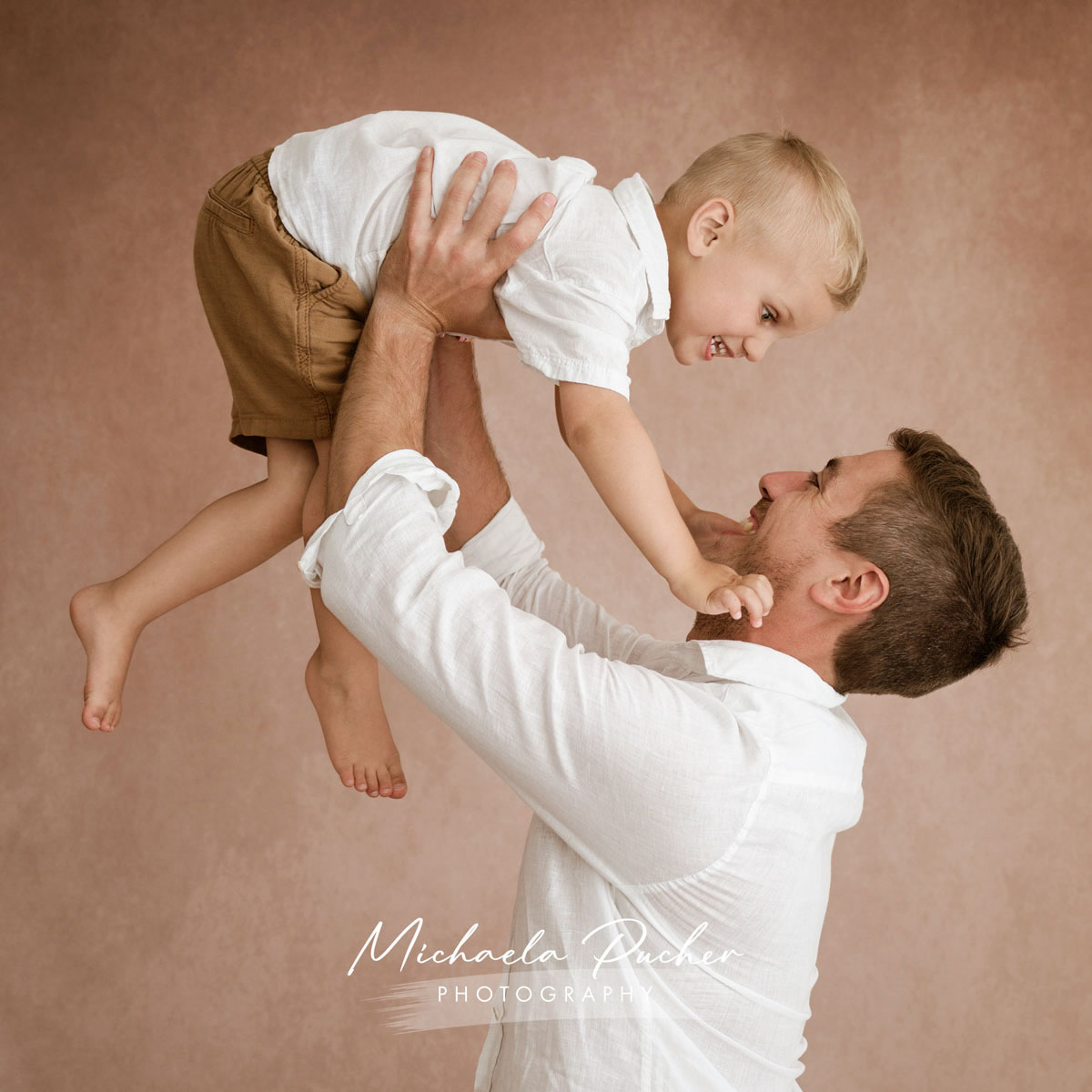 Ein Vater hält sein lachendes Kleinkind kopfüber hoch, beide schauen sich fröhlich an, vor weichem roséfarbenem Hintergrund.